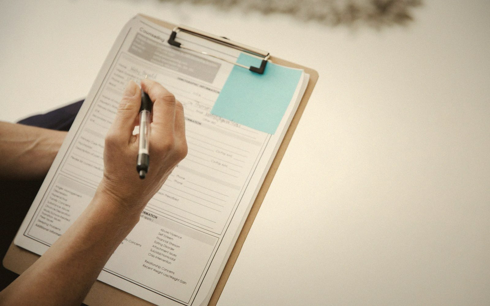 A hand fills out a printed intake form on a clipboard with a sticky note attached