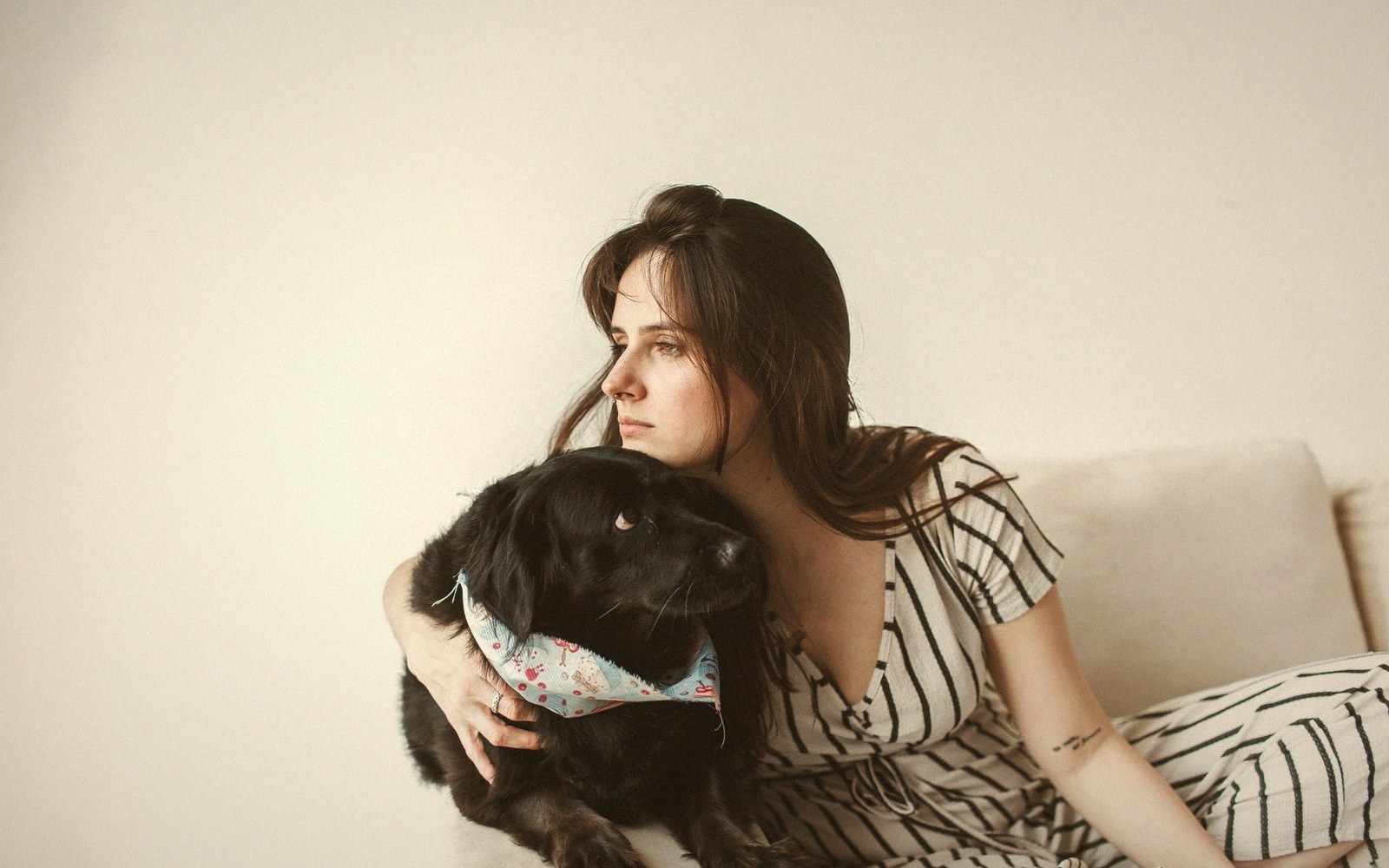 Woman sitting on a couch embraces a black dog wearing a floral bandana